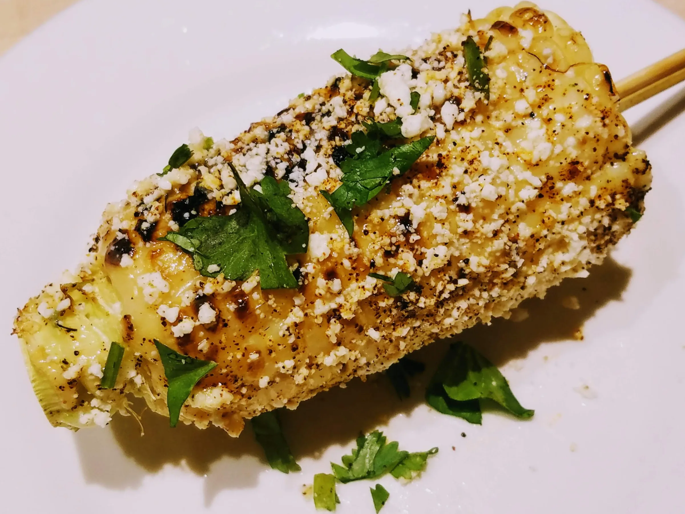 Grilled corn on a skewer, topped with cheese, spices, and cilantro on a white plate.