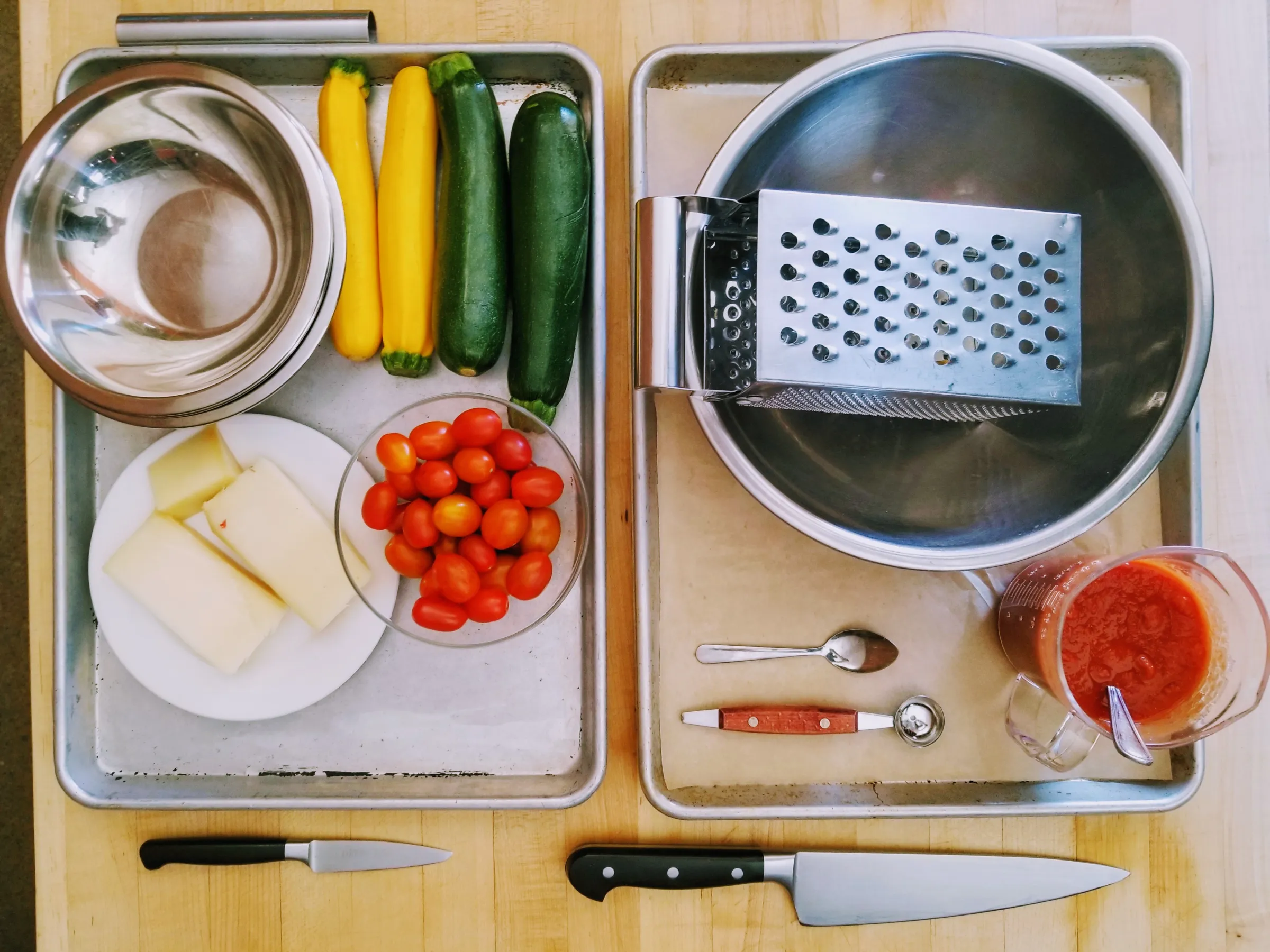 Ingredients and tools for cooking: cheese, tomatoes, zucchini, bowls, graters, knives, and sauce on trays.