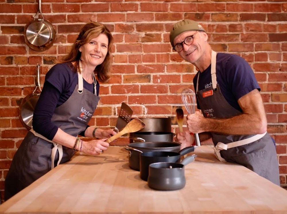 Two people in aprons smiling and holding kitchen utensils in a brick-walled kitchen.